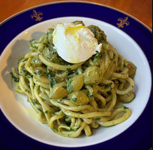A plate of pasta topped with a poached egg and a green pesto sauce, garnished with herbs.