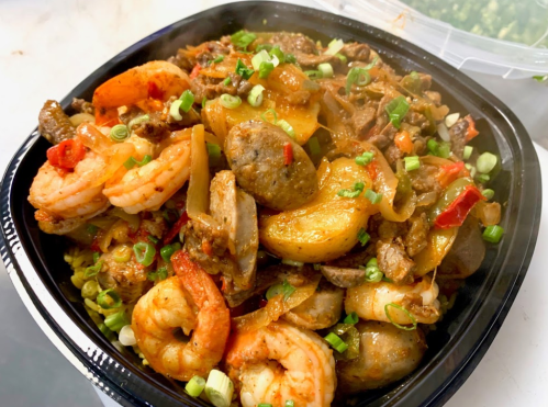 A black takeout container filled with a mix of shrimp, meats, and vegetables, garnished with green onions.