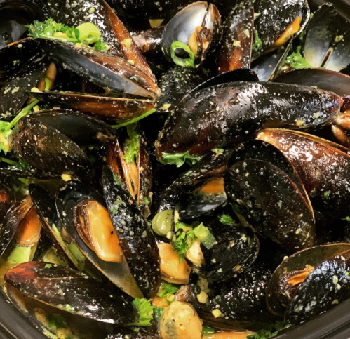 A close-up of cooked mussels garnished with herbs and spices in a dark bowl.