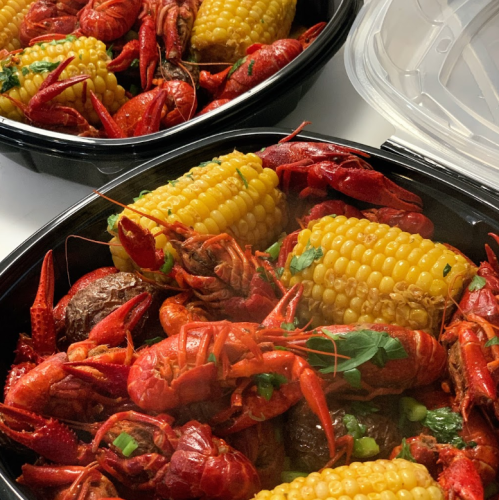A close-up of two containers filled with crawfish, corn, and potatoes, garnished with herbs.