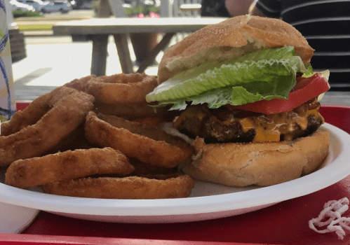 A cheeseburger with lettuce and tomato on a bun, served with a side of crispy onion rings on a white plate.