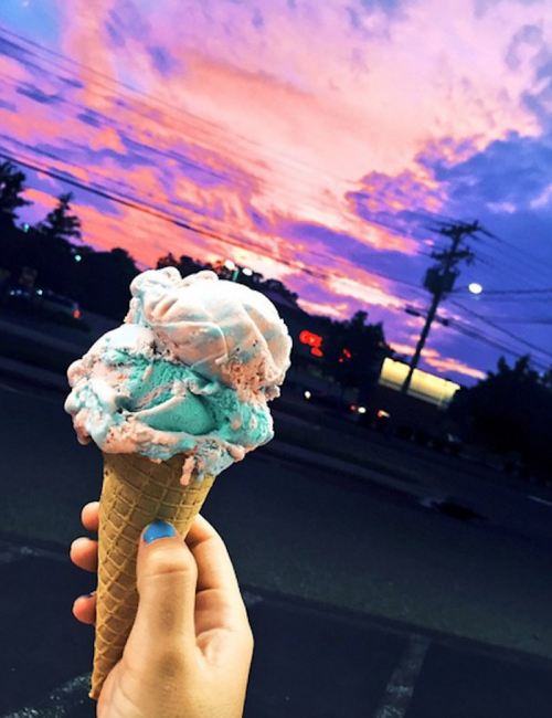 A hand holds a colorful ice cream cone against a vibrant sunset sky with pink and purple hues.
