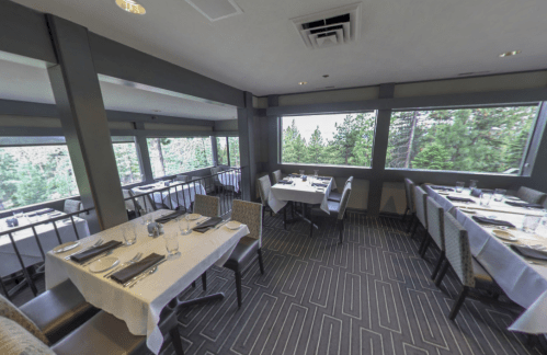 A cozy restaurant interior with tables set for dining, featuring large windows overlooking a forested landscape.