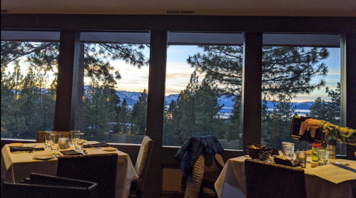 A cozy restaurant with tables set for dining, featuring large windows showcasing a scenic view of trees and mountains at sunset.