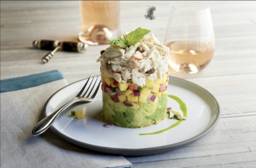 A layered dish of avocado, mango, and crab topped with a mint leaf, served on a plate with a fork and a glass of rosé.