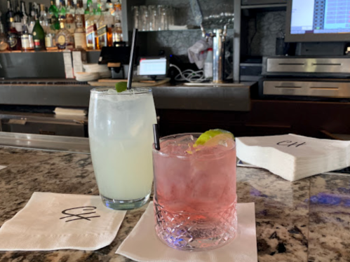 Two cocktails on a bar counter: a light-colored drink with a lime and a pink drink with a lime wedge, on napkins.