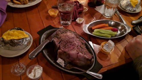 A sizzling platter of seasoned meat with a baked potato, vegetables, and condiments on a wooden table.