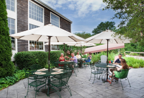 Outdoor dining area with tables and umbrellas, people enjoying meals, surrounded by greenery and a rustic building.