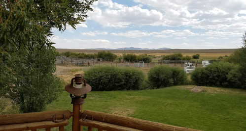 A scenic view from a wooden deck, featuring green grass, shrubs, and distant mountains under a partly cloudy sky.