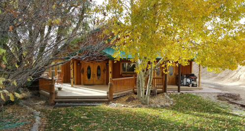 A wooden house surrounded by autumn trees with yellow leaves, featuring a spacious porch and a grassy area.