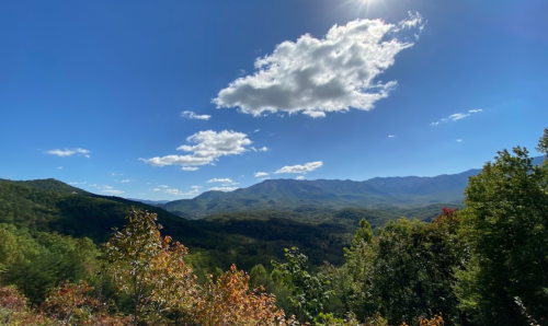 A scenic view of mountains under a blue sky with fluffy clouds, surrounded by lush green trees and autumn foliage.