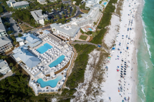 Aerial view of a beach resort with pools, surrounded by greenery and a sandy beach with people and umbrellas.