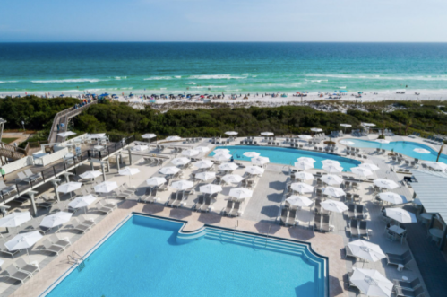 Aerial view of a resort with multiple pools and umbrellas, overlooking a sandy beach and turquoise ocean.