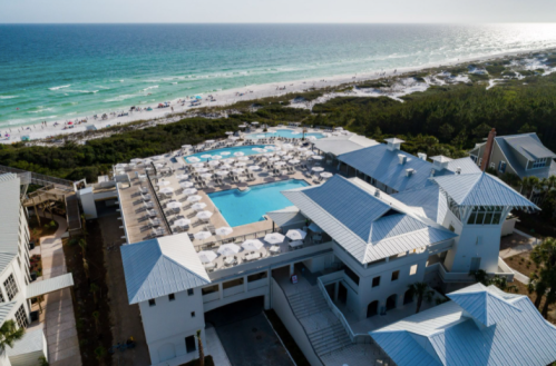 Aerial view of a resort with a large pool, surrounded by beach, ocean, and lush greenery.