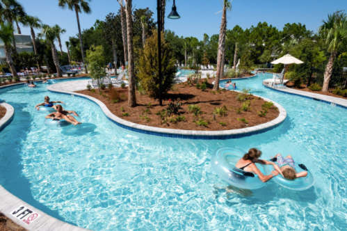 People float on inner tubes in a winding pool surrounded by palm trees and greenery on a sunny day.