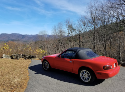 A red convertible car parked on a scenic overlook with mountains and bare trees in the background.