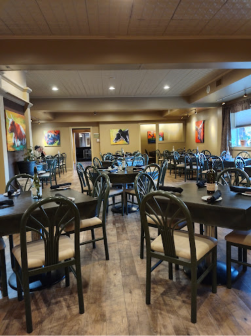 A spacious restaurant interior with wooden floors, green chairs, and colorful artwork on the walls. Tables are set for dining.