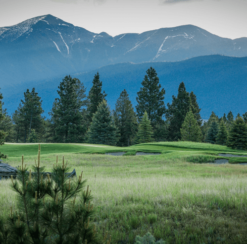 A scenic view of a golf course surrounded by lush greenery and mountains in the background under a cloudy sky.