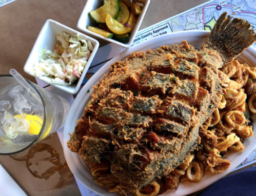A large fried fish served on a platter with fried calamari, coleslaw, and sautéed vegetables, alongside a drink.