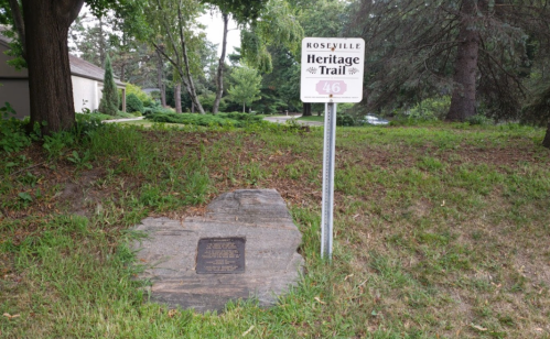 A sign for the Roseville Heritage Trail next to a stone plaque on a grassy area with trees in the background.
