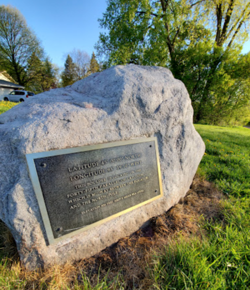 A stone marker with a plaque detailing latitude and longitude coordinates, surrounded by grass and trees.
