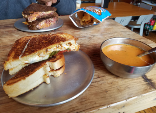 A grilled cheese sandwich with a bite taken out, served with a bowl of tomato soup on a wooden table.