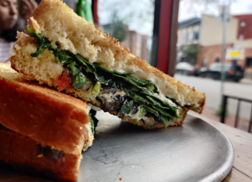 A close-up of a toasted sandwich filled with greens and tomatoes, resting on a plate with a street view in the background.
