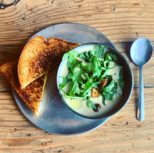 A bowl of creamy soup topped with arugula, accompanied by a toasted cheese sandwich on a metal plate.