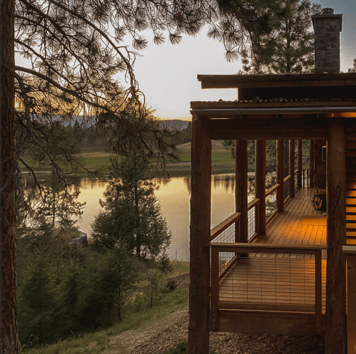 A serene lakeside view at sunset, featuring a wooden deck and trees reflecting in the calm water.