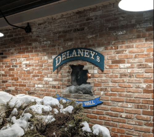 A brick wall with a sign reading "Delaney's" and a hole in the wall, surrounded by snow and greenery.