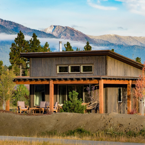 A modern wooden house surrounded by trees and mountains, with a clear sky in the background.