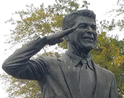 A bronze statue of a smiling man in a suit, saluting with one hand against a backdrop of greenery.
