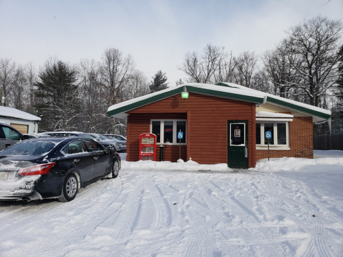 A snow-covered building with a green roof, parking lot, and accessible entrance, surrounded by trees in winter.
