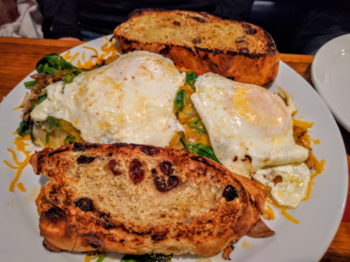 A plate of sautéed vegetables topped with two fried eggs and served with toasted raisin bread.
