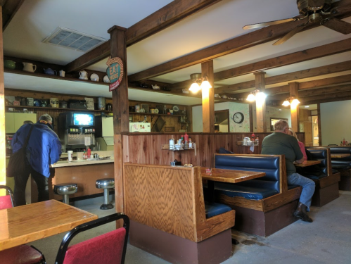 A cozy diner interior with wooden beams, booths, and a vintage soda machine. Two patrons are seated and standing.