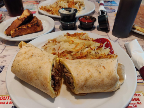 A plate with two burritos, crispy hash browns, ketchup, and two dipping sauces in small cups.