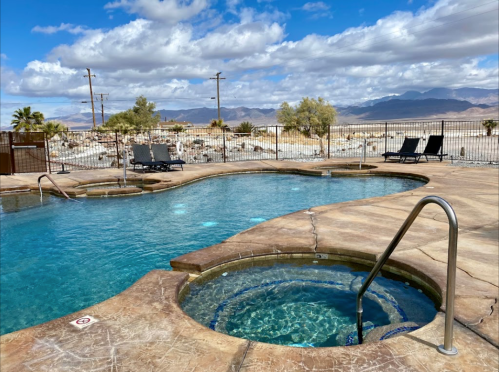 A scenic pool area with a hot tub, surrounded by desert landscape and mountains under a partly cloudy sky.