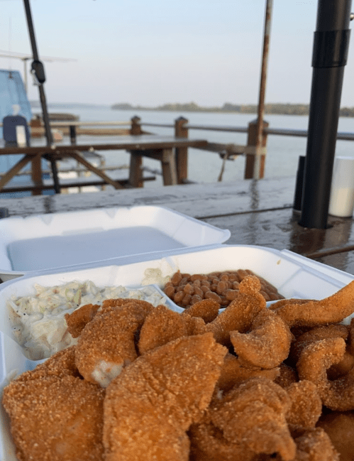 A takeout box with fried seafood, coleslaw, and beans, set on a wooden table by a river at sunset.