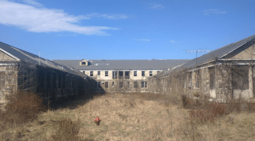 Abandoned building complex with overgrown grass and a clear blue sky.