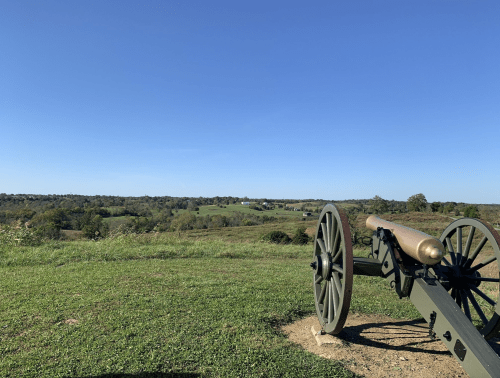 A historic cannon on a grassy hill, overlooking a vast landscape under a clear blue sky.