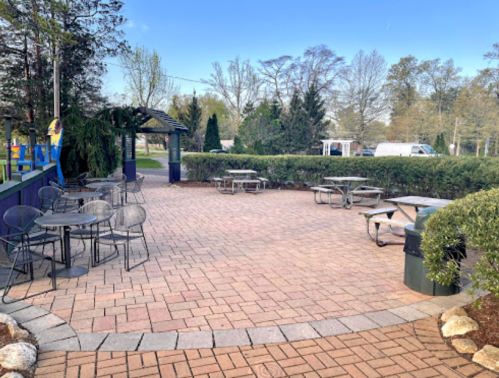 Outdoor seating area with tables and chairs, surrounded by greenery and a paved walkway under a clear blue sky.