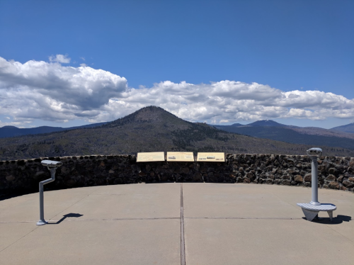 View from a lookout point featuring mountains, a stone wall, and informational plaques under a blue sky with clouds.