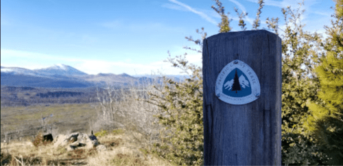 A wooden post with a trail marker stands against a backdrop of mountains and clear blue sky.