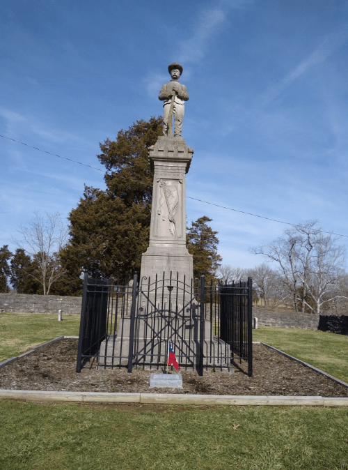 A tall statue of a soldier on a pedestal, surrounded by a black iron fence, set against a clear blue sky.
