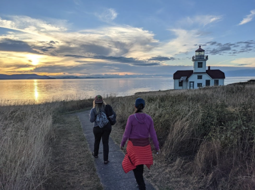 Two people walk along a path towards a lighthouse at sunset, with calm waters and a colorful sky in the background.