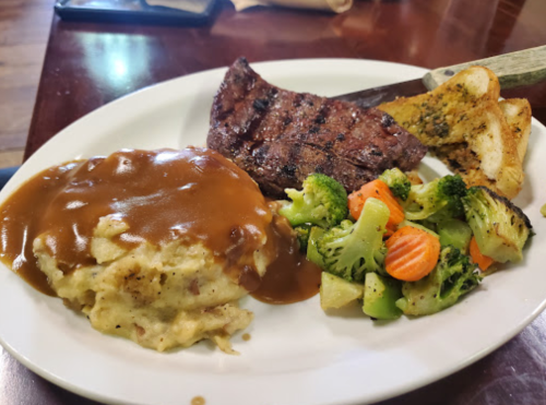 A plate with grilled steak, mashed potatoes with gravy, mixed vegetables, and garlic bread.