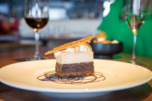 A layered dessert with brownie, mousse, and a crispy topping, served on a white plate with wine glasses in the background.
