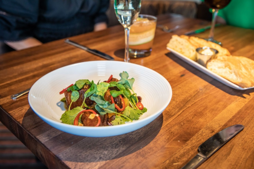 A bowl of salad with greens, red peppers, and meatballs, accompanied by drinks and bread on a wooden table.