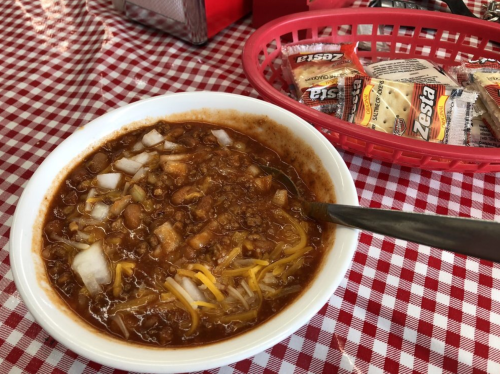 A bowl of chili topped with cheese and onions, served on a checkered tablecloth, with condiments in the background.
