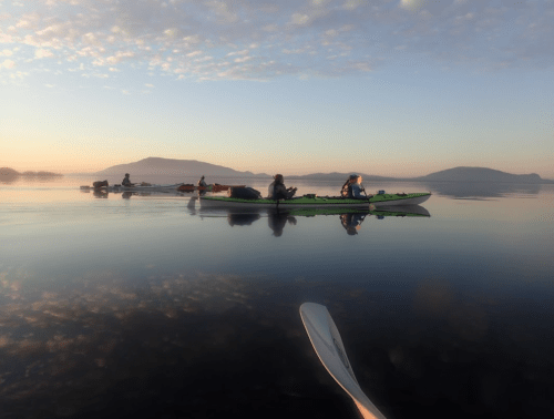 A serene early morning scene of kayakers paddling on calm water with misty mountains in the background.